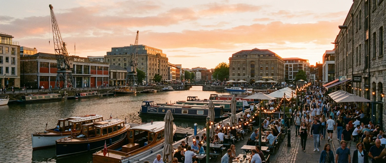 Panoramic view of Bristol harbourside at golden hour with waterfront restaurants, boats moored in the harbor, historic buildings, vibrant atmosphere, people dining outdoors, warm evening lighting